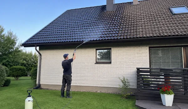 Katuse pehmepesu ja tõrjetöötlus, meister pritsib pika varrega pesulahust tumedale kivikatusele, murul pritsipaak, pritsi vars on süsinikkiust ja nii kerge, et meister juhib seda ühe käega.