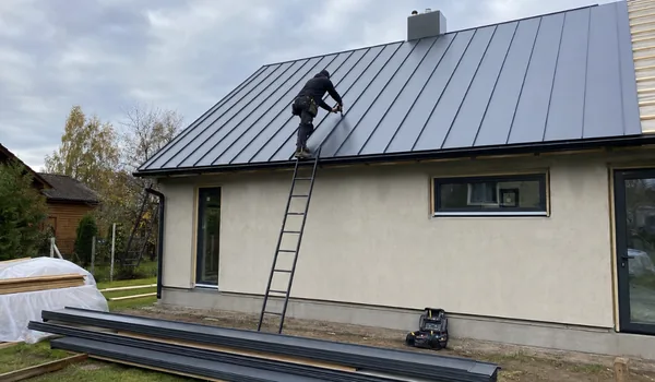 Plekk-katuse paigaldus, meister kinnitab heleda krohvitud maja katusele tumedat valtsplekki, maas plekipaanid, vihmamüra summutab aluskate, mitte pleki paksus.